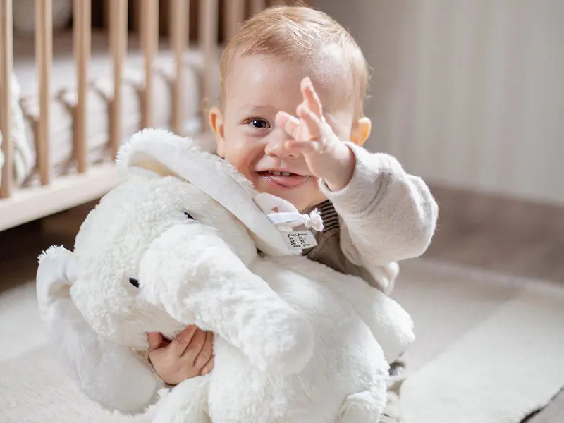 Baby met knuffel in de hand die zwaait naar de fotograaf, mooi houten kinderbedje in de achtergrond.