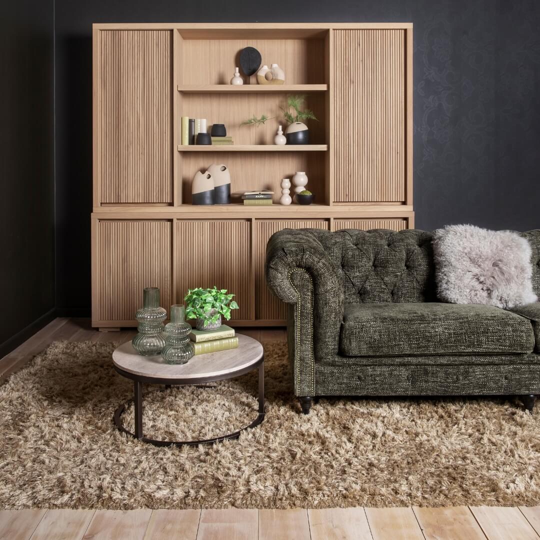 Gezellige hoek met groene bank en houten opbergkast, inclusief decoratieve accessoires en een shaggy vloerkleed. Perfect voor een warme herfstsfeer met de 'Cocoon Deals'.